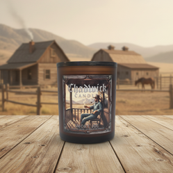 Candle in a decorative holder with a cowboy design on a wooden table in front of a rustic landscape.