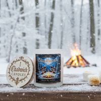 Candle with packaging and wooden coaster on a snowy surface with a campfire in the background